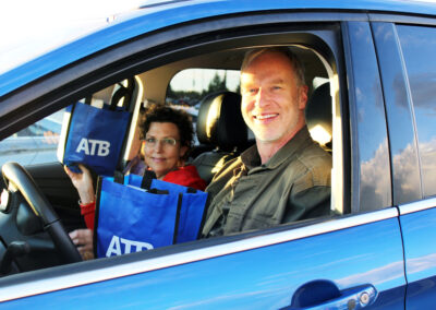 Two people sitting in the front seats of a blue car. They are smiling and holding blue reusable bags that say "ATB"