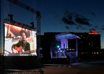 Large outdoor screen with Barney Bentall. He is wearing a hat and playing a guitar. In the background is the River Cree Hotel with a red neon sign that says "River Cree"