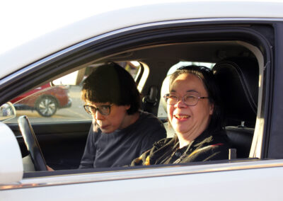 Two people sitting in the front seats of a silver car with the window rolled down, smiling at the camera