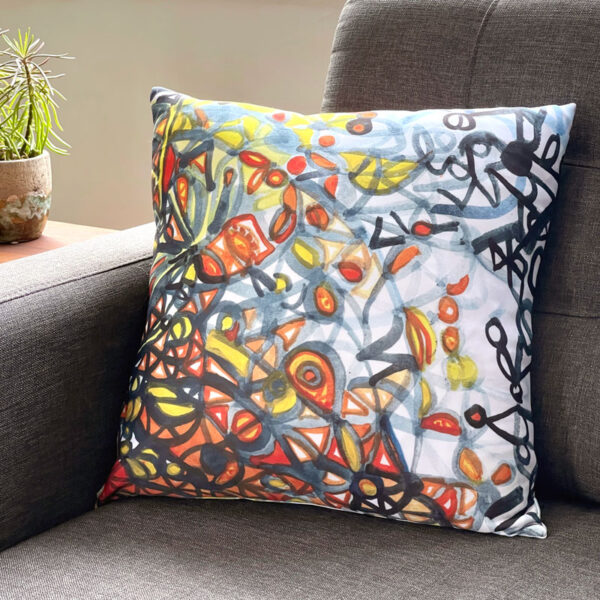 Square pillow featuring an orange, blue, and yellow painting by Uli Rossier on a grey couch