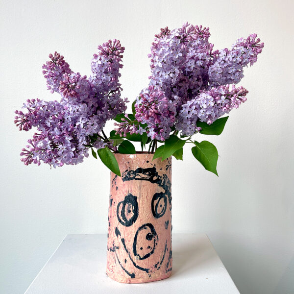 Pink ceramic vase with a smiley face painted on it. The vase is full of lilacs
