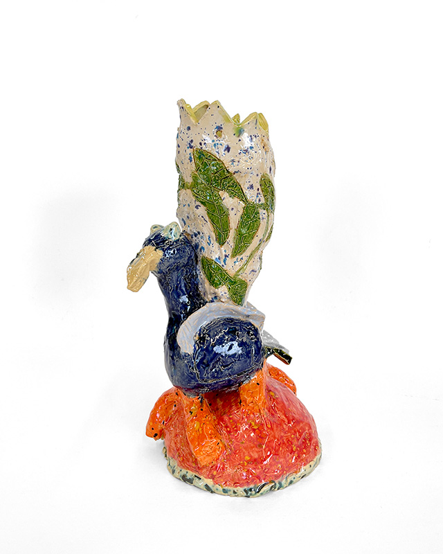 Carrie Wheeler Bird Vase sm Ceramic vase with an orange base and a blue bird. The body if the vase is white with green leaves on it.